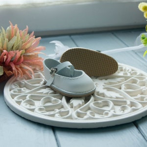 May include: A pair of white leather baby shoes with a silver buckle. The shoes are on a white decorative plate with a floral design.