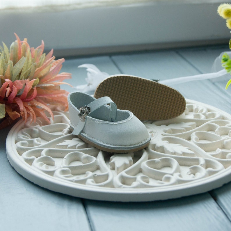 May include: A pair of white leather baby shoes with a silver buckle. The shoes are on a white decorative plate with a floral design.