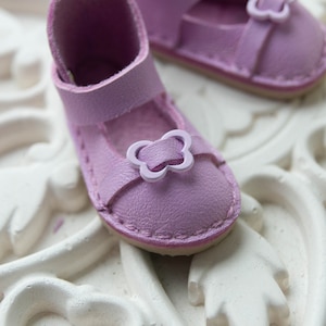 May include: A pair of small, lavender-colored Mary Jane style shoes. Each shoe has a strap with a light purple flower-shaped buckle. The shoes are made of a soft material and have stitched detailing around the edges.