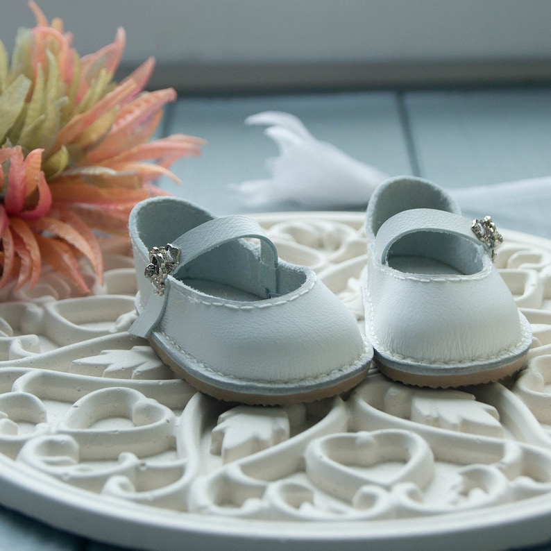 May include: Two white leather baby shoes with a strap and a small silver buckle. The shoes are on a white ornate surface.