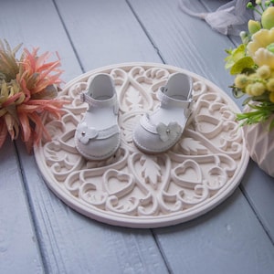 May include: A pair of white leather baby shoes with a flower detail on a white decorative surface.