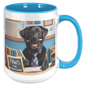 May include: White ceramic coffee mug with a blue rim and handle. The mug features a black Labrador Retriever wearing glasses and a tie, sitting at a desk with a book and a chalkboard that says "Paws for Knowledge".