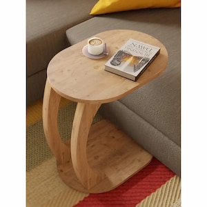 May include: A light-colored wooden side table with a curved base. A small cup of coffee sits on a saucer, and a book titled "Running Toward Dawn" rests on the table's surface. The table is positioned next to a gray sofa.