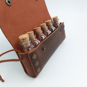 Puede incluir: Una bolsa de cuero marr&oacute;n con una solapa y una correa. La bolsa est&aacute; abierta y contiene seis peque&ntilde;as botellas de vidrio con tapones de corcho.