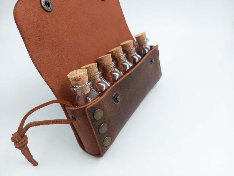Puede incluir: Una bolsa de cuero marr&oacute;n con una solapa y una correa. La bolsa est&aacute; abierta y contiene seis peque&ntilde;as botellas de vidrio con tapones de corcho.