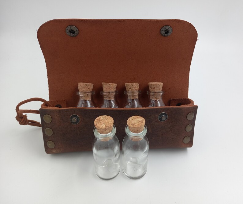 Puede incluir: Una bolsa de cuero marr&oacute;n con seis peque&ntilde;as botellas de vidrio con tapones de corcho. La bolsa tiene una solapa que se abre para revelar las botellas.