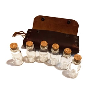 Puede incluir: Un juego de seis peque&ntilde;as botellas de vidrio con tapones de corcho, dispuestas en fila sobre una superficie blanca. Las botellas son transparentes y est&aacute;n vac&iacute;as. Una caja de cuero marr&oacute;n con una solapa est&aacute; abierta y se encuentra detr&aacute;s de las botellas.
