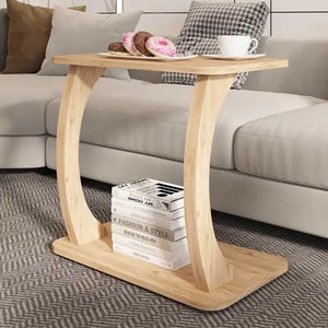 Pode incluir: Uma mesa lateral de madeira clara com um tampo retangular e pernas curvas. A mesa tem uma prateleira inferior com livros. Um prato de donuts e uma xícara de café estão em cima. A mesa tem 71 cm de comprimento, 36 cm de largura e 61 cm de altura.