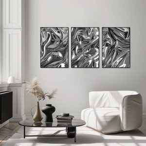 Wohnzimmerszene mit einem Sessel und einem niedrigen Tisch, der mit Büchern und Vasen dekoriert ist. An der Wand sieht man 3 schwarze Rahmen, die eine abstrakte und unique Plakat-Serie zeigen. Die Motive zeigen verschiedene Alu-Strukturen.