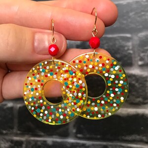 May include: A pair of gold-toned hoop earrings with a clear, yellow-tinted center. The hoops are decorated with colorful dots in red, yellow, blue, and white. Each earring has a small red bead and gold-colored hook.