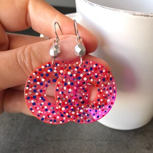 May include: A pair of pink hoop earrings with a speckled design in red, white, and blue. The earrings have silver-colored ear wires and a faceted bead detail. The earrings are a fun and colorful accessory.