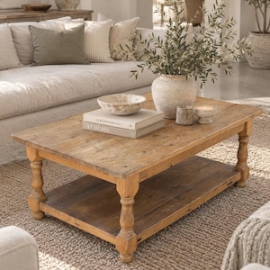 May include: A rectangular wooden coffee table with four turned legs and a lower shelf. On the table sits a stone bowl, two stacked books titled The Kinfolk Home, a textured ceramic vase with olive branches, and two small stone candle holders.