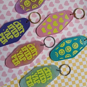 May include: Five colorful keychains with different designs. Each keychain is shaped like a motel key and features the text "CERTIFIED YAP PER" in yellow. The keychains feature a smiley face pattern, a heart pattern, a turquoise blue color, a pink color, and a purple color.