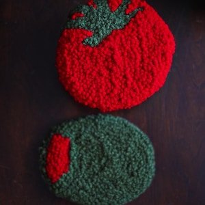 May include: Two handmade, textured coasters. One is a red tomato shape with a green stem. The other is a green olive shape with a red accent. These coasters are designed to protect surfaces from drink condensation.