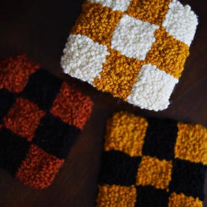 May include: Three square, hand-stitched coasters with a checkerboard pattern. One coaster features a white and gold pattern, another has a black and gold pattern, and the third has a black and rust-colored pattern. The coasters are made of a textured, looped yarn.
