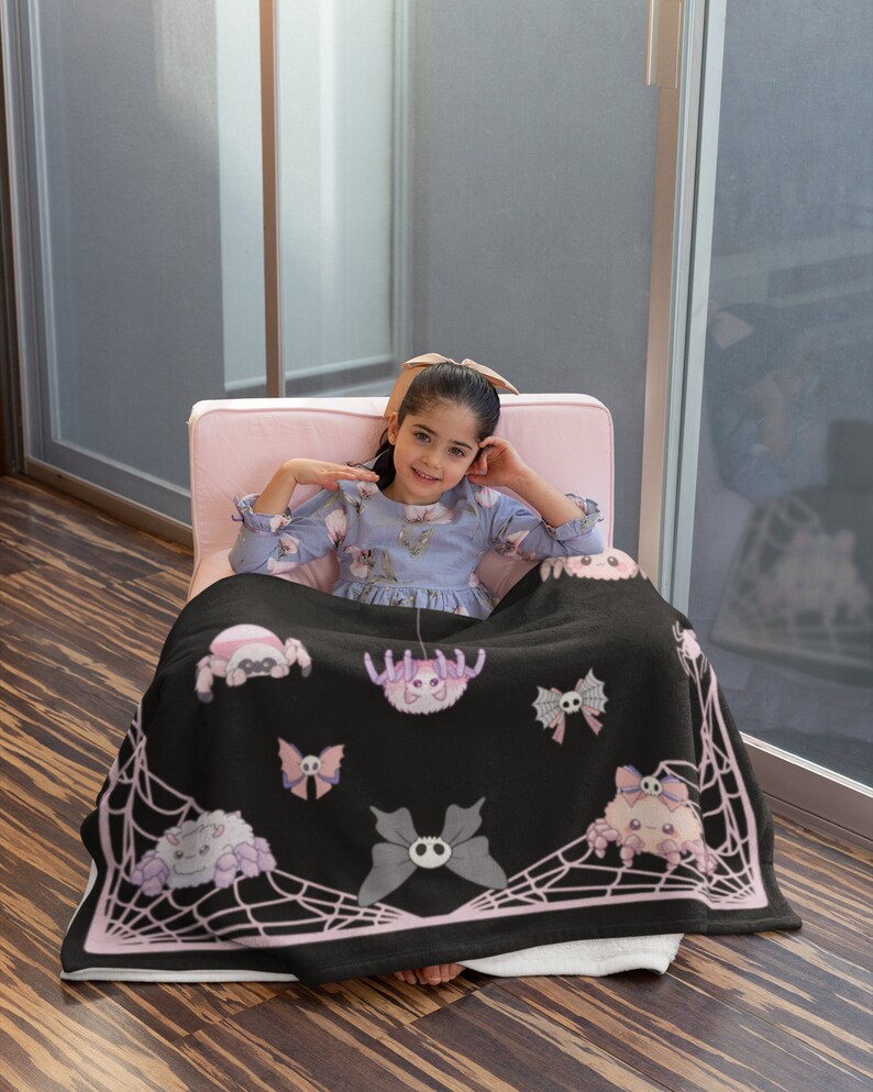 May include: A black fleece throw blanket with a pink spiderweb design and pink cartoon spiders. The blanket features a skull and crossbones design on the butterflies and spiders.