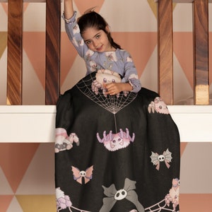 May include: A black and pink blanket with a spider web design and cute spider illustrations. The blanket is draped over a railing and a young girl is sitting on it.