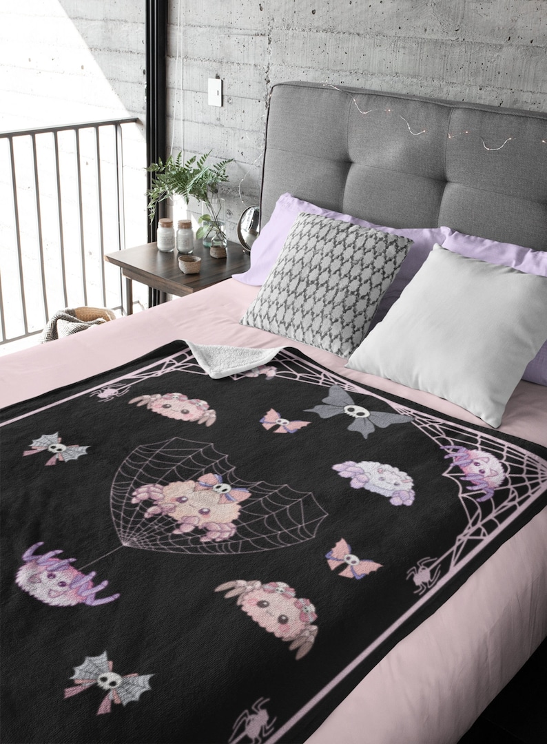 May include: A black and pink fleece throw blanket with a spiderweb design and cute cartoon spiders. The spiders are wearing bows and have skulls on their heads.