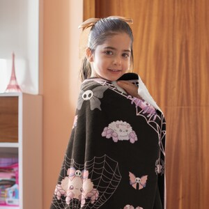 May include: A black fleece blanket with a pink and white spider web design. The blanket features cute cartoon spiders with bows and skulls.