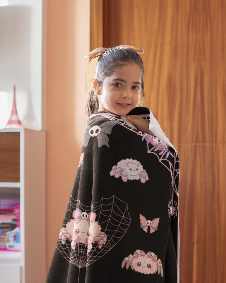 May include: A black fleece blanket with a pink and white spider web design. The blanket features cute cartoon spiders with bows and skulls.