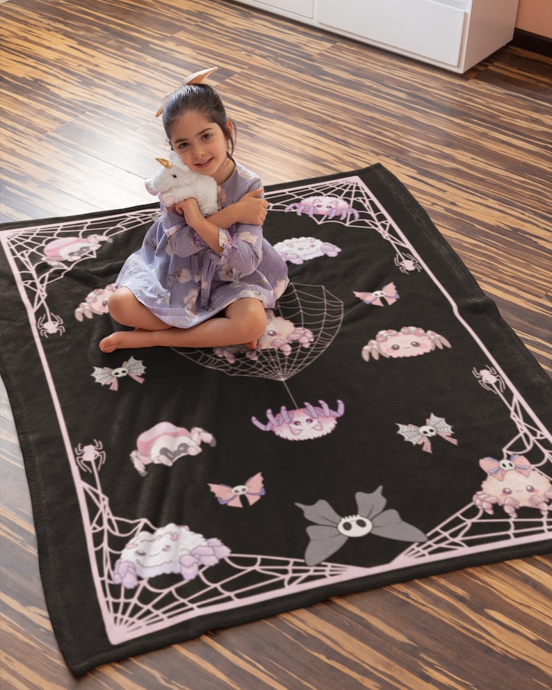 May include: A black and pink fleece blanket with a spiderweb design and cute cartoon spiders. The blanket features a pink spiderweb pattern with various cartoon spiders in pink, white, and grey.