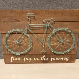 May include: Wooden wall art featuring a bicycle design crafted with string and nails. The bicycle is light blue against a brown wooden background. The phrase "find joy in the journey" is written in white script.
