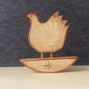 May include: Wooden chicken figurine with a cream-colored body, red accents, and a yellow beak. The chicken stands on a curved base, and the design includes a stylized wing and tail. A charming piece of folk art.