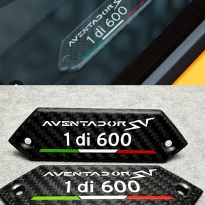 Puede incluir: Una placa de fibra de carbono negra con texto blanco que dice "AVENTADOR SV 1 di 600". La placa tiene una franja verde, blanca y roja en la parte inferior, que se asemeja a la bandera italiana.