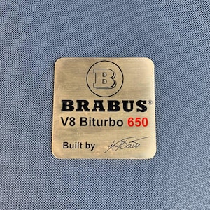 May include: A rectangular, brushed metal emblem with the text "BRABUS" in black, "V8 Biturbo 650" in red, and "Built by" with a signature. The emblem features a circular logo with a "B" inside.