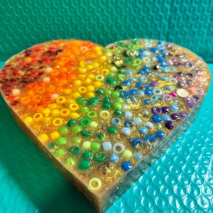 May include: A heart-shaped resin art piece filled with colorful beads. The beads are arranged in a rainbow pattern, transitioning from red to blue. The resin has a golden shimmer, and the edges are a light brown.