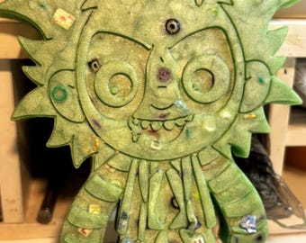 May include: A light green, semi-translucent, flat-backed figurine featuring a stylized character with spiky hair, large circular eyes, and a wide grin. The surface is embedded with small colorful confetti, beads, and letter tiles.