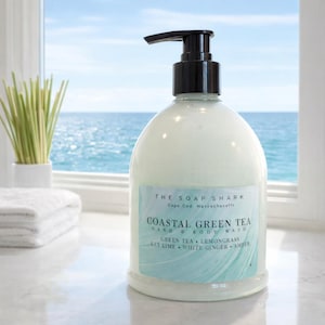 Puede incluir: Una botella blanca en forma de pera de jabón para manos y cuerpo con una bomba negra. La etiqueta dice "Coastal Green Tea" con los ingredientes enumerados. El fondo muestra una ventana con vista al océano y un jarrón pequeño.