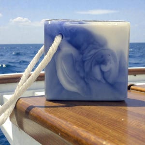 May include: A square soap bar with a blue and white marbled design and a white rope attached. The soap is resting on a wooden surface with the ocean and sky in the background.