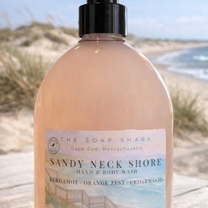 May include: A clear, peach-colored hand and body wash bottle with a black pump. The label reads "Sandy Neck Shore" and lists bergamot, orange zest, and cedarwood. The background shows a beach scene.
