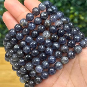 May include: A collection of dark blue, round gemstone beads. The beads are smooth and polished, with variations in color and texture. The beads are strung together, forming a bracelet or necklace. The gemstones are approximately 0.3 inches in diameter.