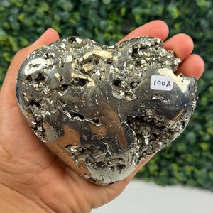 May include: A heart-shaped pyrite crystal, held in a hand, with a metallic, reflective surface. The crystal has a rough, textured appearance with a label that reads "100A". The background is a blurred green.