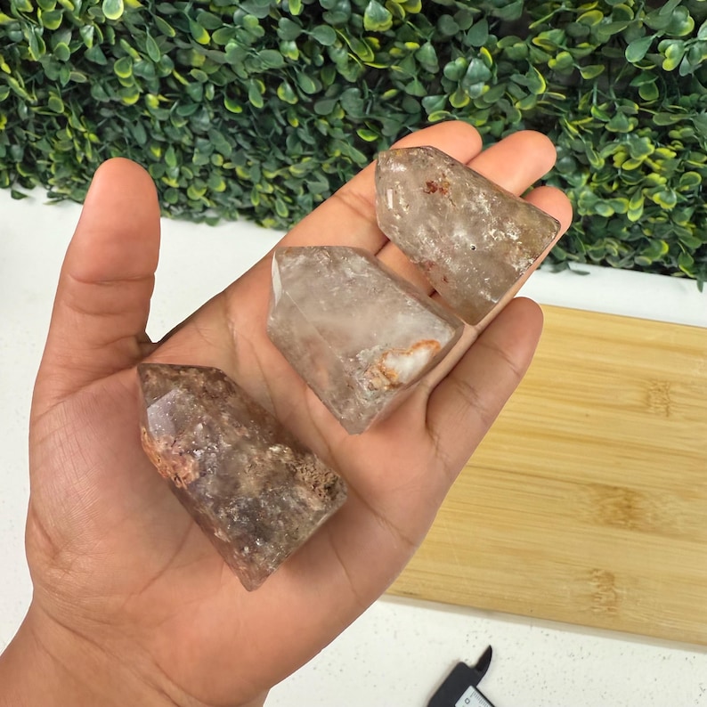 May include: Three clear quartz crystal points with brown and orange inclusions are held in a hand. The crystals have a faceted shape and varying sizes. The background features green foliage and a wooden cutting board.