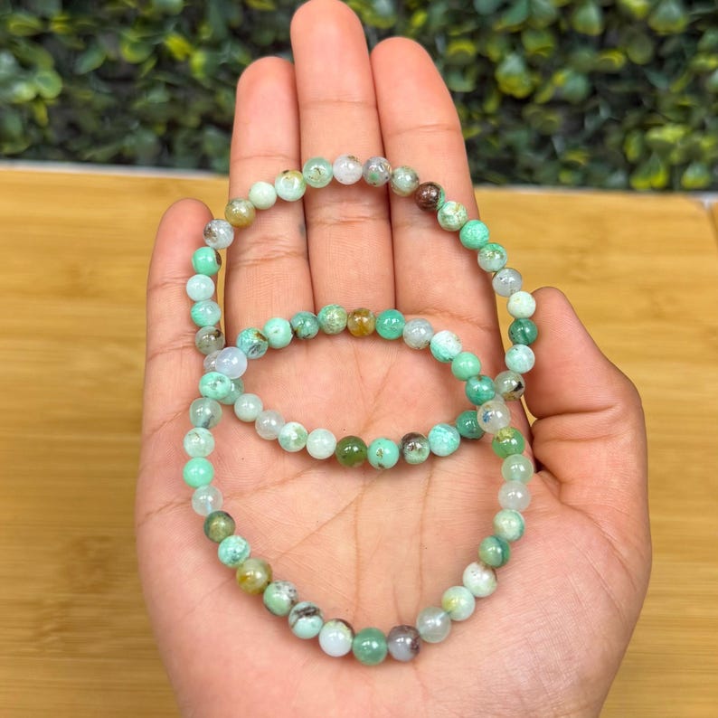 May include: Two bracelets made of small, round, light green and white beads. The beads have a natural, speckled appearance. The bracelets are held in a hand, showcasing their delicate design and color variations.