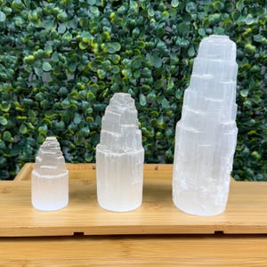 May include: Three white selenite crystal towers of varying sizes are displayed on a wooden tray. The crystals have a layered, textured appearance, with a natural, translucent quality. The background features a green, leafy wall.