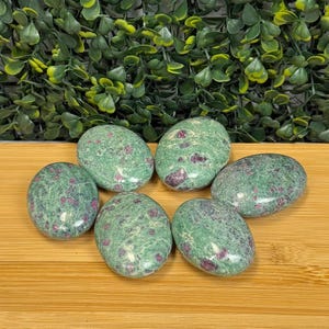 May include: Six oval-shaped stones with a green base color and red inclusions are arranged on a wooden surface. The stones are smooth and polished, with a speckled appearance. Green foliage is in the background.
