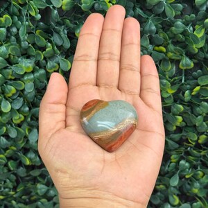 Puede incluir: Una piedra en forma de coraz&oacute;n con una superficie pulida. La piedra presenta bandas de tonos rojos, marrones y verde azulado. La piedra se sostiene en una mano abierta sobre un fondo de follaje verde.