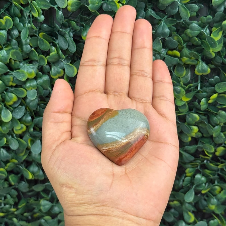 Puede incluir: Una piedra en forma de coraz&oacute;n con una superficie pulida. La piedra presenta bandas de tonos rojos, marrones y verde azulado. La piedra se sostiene en una mano abierta sobre un fondo de follaje verde.