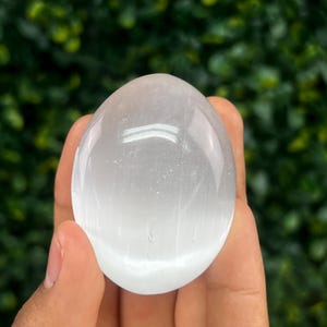 Pode incluir: Cristal de selenite liso e oval, segurado em uma m&atilde;o. A pedra transl&uacute;cida tem uma cor branca perolada e uma superf&iacute;cie polida. O fundo &eacute; verde desfocado, sugerindo folhagem. O cristal tem aproximadamente 7.6 cm de comprimento.