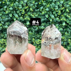 May include: Two clear quartz crystal points with inclusions, held in hands. The crystals have a natural, pointed shape with areas of grey and brown within the clear quartz. A black square with the text "#4" is in the background.