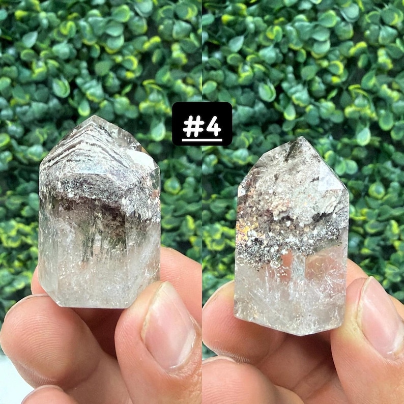May include: Two clear quartz crystal points with inclusions, held in hands. The crystals have a natural, pointed shape with areas of grey and brown within the clear quartz. A black square with the text "#4" is in the background.