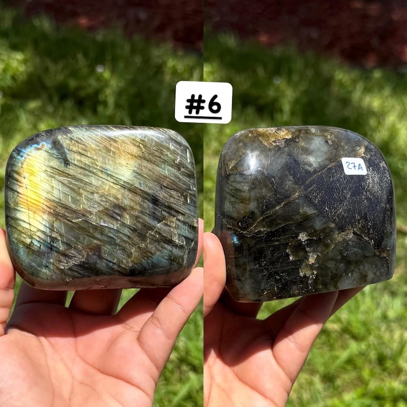 May include: Two polished labradorite stones with iridescent flashes of yellow, blue, and grey. The left stone is labeled #6, and the right stone features a small white sticker with the text 27A. Both stones have a smooth, rounded surface.