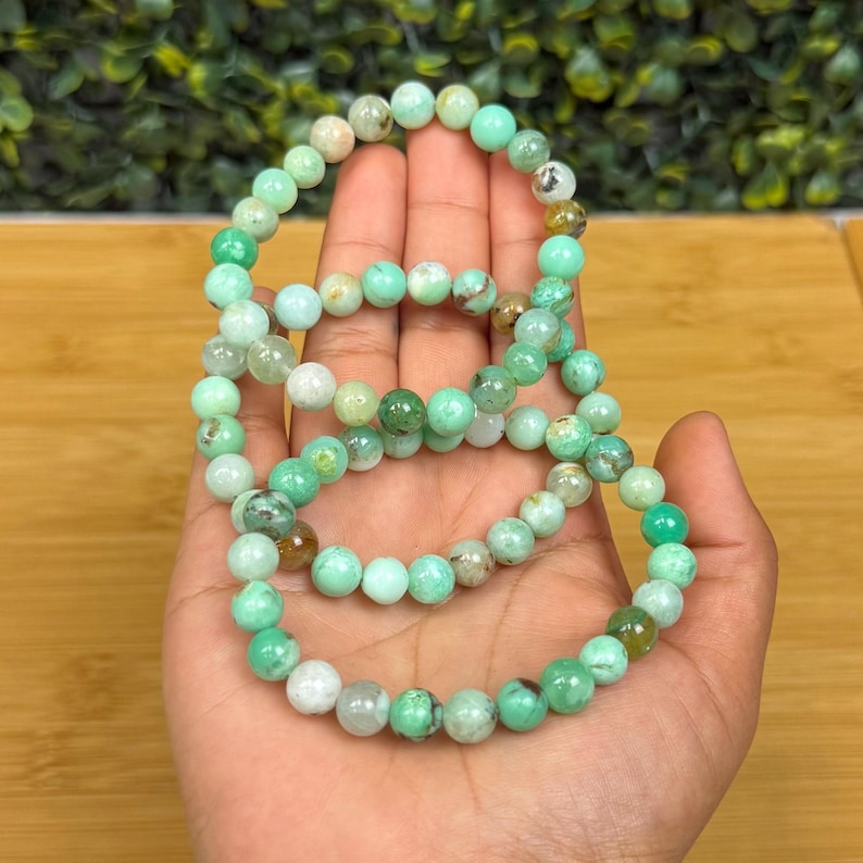 May include: Three beaded bracelets with green and white round stones. The bracelets are held in a hand, showcasing the varying shades of green and white in the stones. The background is a mix of green foliage and a wooden surface.