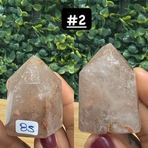 May include: Two clear quartz crystal points with a light brown hue and internal inclusions. One has a sticker with the number "85". The background is a green leafy pattern, and a black square with the number "#2" is in the background.