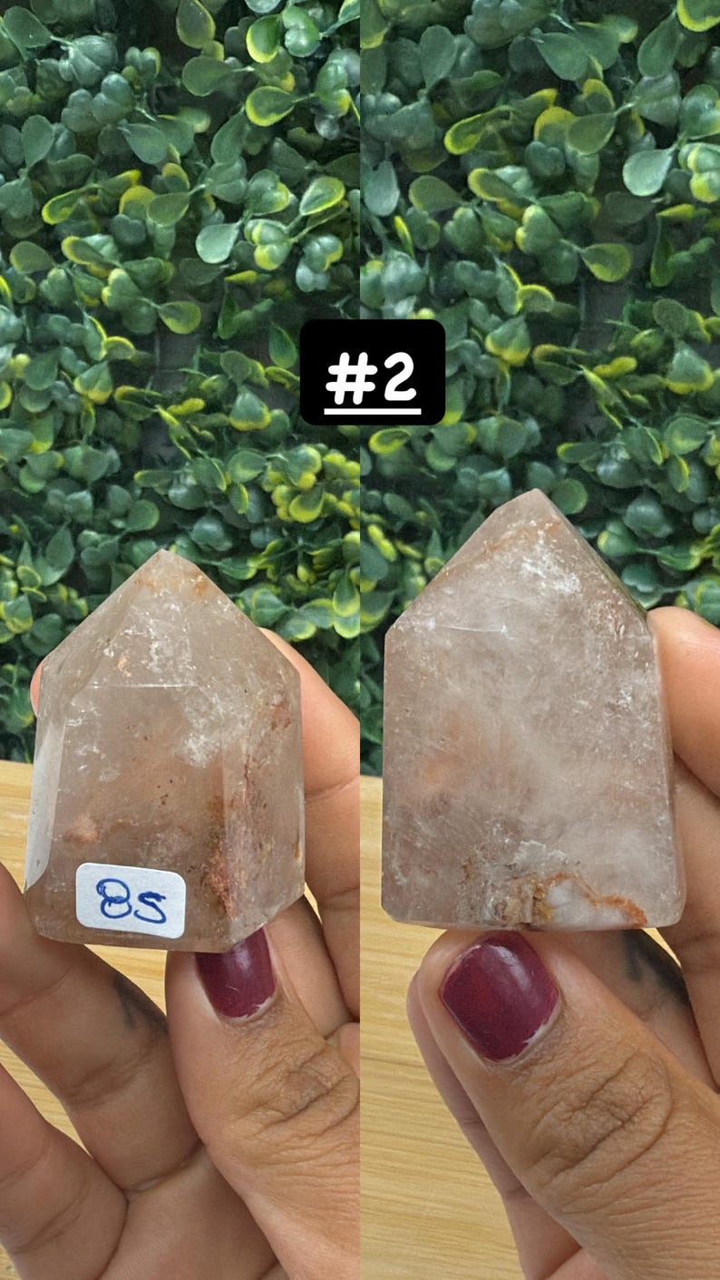 May include: Two clear quartz crystal points with a light brown hue and internal inclusions. One has a sticker with the number "85". The background is a green leafy pattern, and a black square with the number "#2" is in the background.
