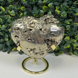 May include: A heart-shaped pyrite specimen with a metallic, silver-gold color. The pyrite is mounted on a gold-colored metal stand. The stand has a circular base and two small supports. The text "100A" is visible on the pyrite. The background features green foliage.